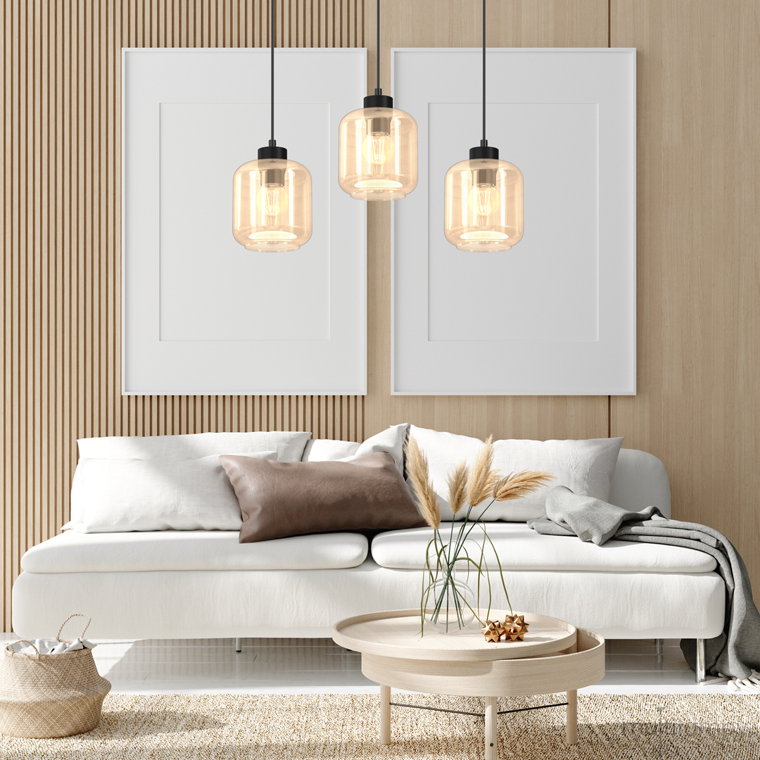 Lampa wisząca 3-punktowa Sombra w kolorze czarnym z bursztynowym kloszem, zawieszona nad jasną sofą, w nowoczesnym wnętrzu z drewnianymi panelami. Widać trzy szklane klosze i elementy dekoracyjne, takie jak stół i poduszki.