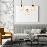 Lampa wisząca Sombra 3-punktowa w kolorze czarnym z bursztynowymi kloszami, zawieszona na czarnych przewodach. Widoczna w nowoczesnym wnętrzu z szarym sofą, czarnymi stolikami kawowymi i żółtym fotelem. Tło z marmurową ścianą i białą płaską powierzchnią.