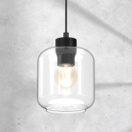 Lampa wisząca Sombra 1-punktowa, czarna z przezroczystym kloszem, o cylindrycznym kształcie. Widoczna żarówka w środku, zawieszona na cienkim przewodzie. Tło w kolorze szarym, betonowym, podkreślające minimalistyczny styl.