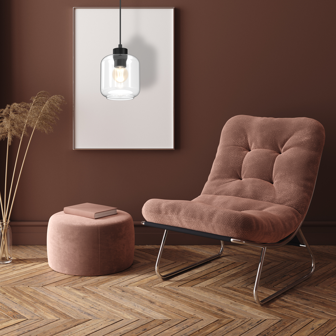 Lampa wisząca Sombra 1-punktowa w kolorze czarnym z transparentnym kloszem, zawieszona nad różowym fotelem w nowoczesnym wnętrzu. Klosz o cylindrycznym kształcie, z widoczną żarówką, wisi na cienkim kabelku. Tło w odcieniu brązu.