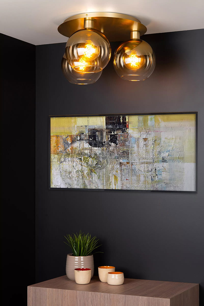 Złota lampa sufitowa Marius z trzema okrągłymi kloszami z przezroczystego szkła, montowana na okrągłej podstawie. Lampa emituje ciepłe światło, wisząca w nowoczesnych wnętrzach. Tło ciemne, z obrazem i dekoracjami na drewnianym stole.