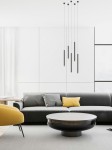 Lampa wisząca Q I Nero/Gold 50 w kolorze czarnym i złotym, składająca się z kilku cienkich, prostokątnych elementów, zawieszona na cienkich przewodach. Produkt umiejscowiony nad okrągłym stolikiem we wnętrzu z szaro-żółtym sofą oraz jasnym tłem.