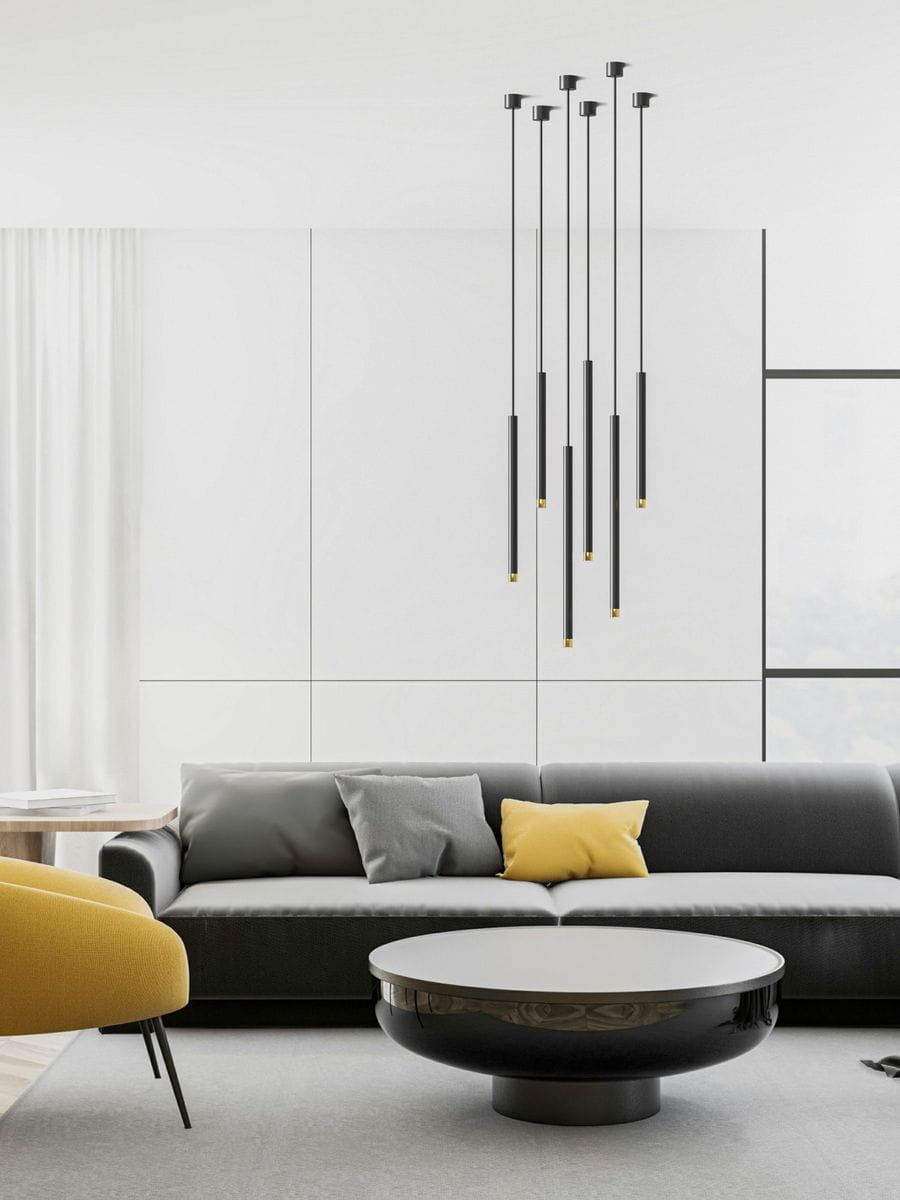 Lampa wisząca Q I Nero/Gold 60 w kolorze czarnym i złotym z sześcioma długimi, smukłymi ramionami. Zawieszona w nowoczesnym wnętrzu z szarym sofą i okrągłym stołem. Tło w jasnych odcieniach, podkreślające elegancki design.
