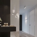 Lampa wisząca 1-punktowa w białym kolorze z kwadratowym kloszem, zawieszona na cienkim przewodzie. Ujęcie z boku, w nowoczesnym wnętrzu, obok czarnej ściany. W tle widoczne meble i drzwi.