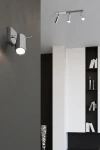 Lampa sufitowa (spot) 3-punktowa w kolorze chromowanym, zamontowana na ścianie. Ujęcie z boku, ukazujące cylindryczny kształt reflektora. W tle minimalistyczne wnętrze z białymi meblami i dekoracjami.