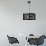 Czarna lampa wisząca Fishnet 3-punktowa z metalowym kloszem w siatkę, zawieszona na łańcuchu, umieszczona nad drewnianym stołem. W tle cztery krzesła i stół, na którym znajdują się talerz i kieliszek.
