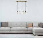Lampa wisząca 4-punktowa Woody w jasnym drewnie z czarnymi oprawkami, zawieszona nad szarym narożnikiem z poduszkami. Widoczne cztery żarówki w klasycznym stylu. Prosta aranżacja na jasnym tle.
