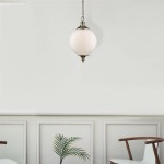 Lampa wisząca Vulcan 1-punktowa w antycznym mosiądzu. Okrągła, matowa kula ze szkła w białym kolorze, zawieszona na łańcuchu. Produkt umieszczony w jasnym wnętrzu z elementami dekoracyjnymi, takimi jak roślina doniczkowa i krzesła.