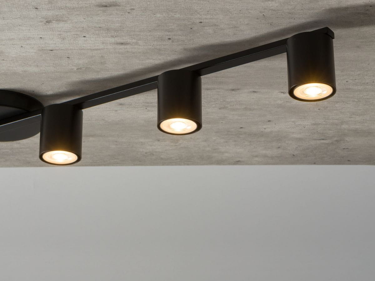 Czarna lampa sufitowa 4-punktowa Sky z trzema cylindrycznymi oprawami, zamontowana na szarej powierzchni. Widoczna struktura sufitu betonowego. Światła emitują ciepłe, białe światło.