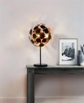 Lampa stołowa 3-punktowa Discus w kolorze czarnym z okrągłymi elementami dekoracyjnymi, umieszczona na ciemnym stole. Źródło światła tworzy efekt świetlny na ścianie. W tle ramka ze zdjęciem oraz wazon z rośliną.