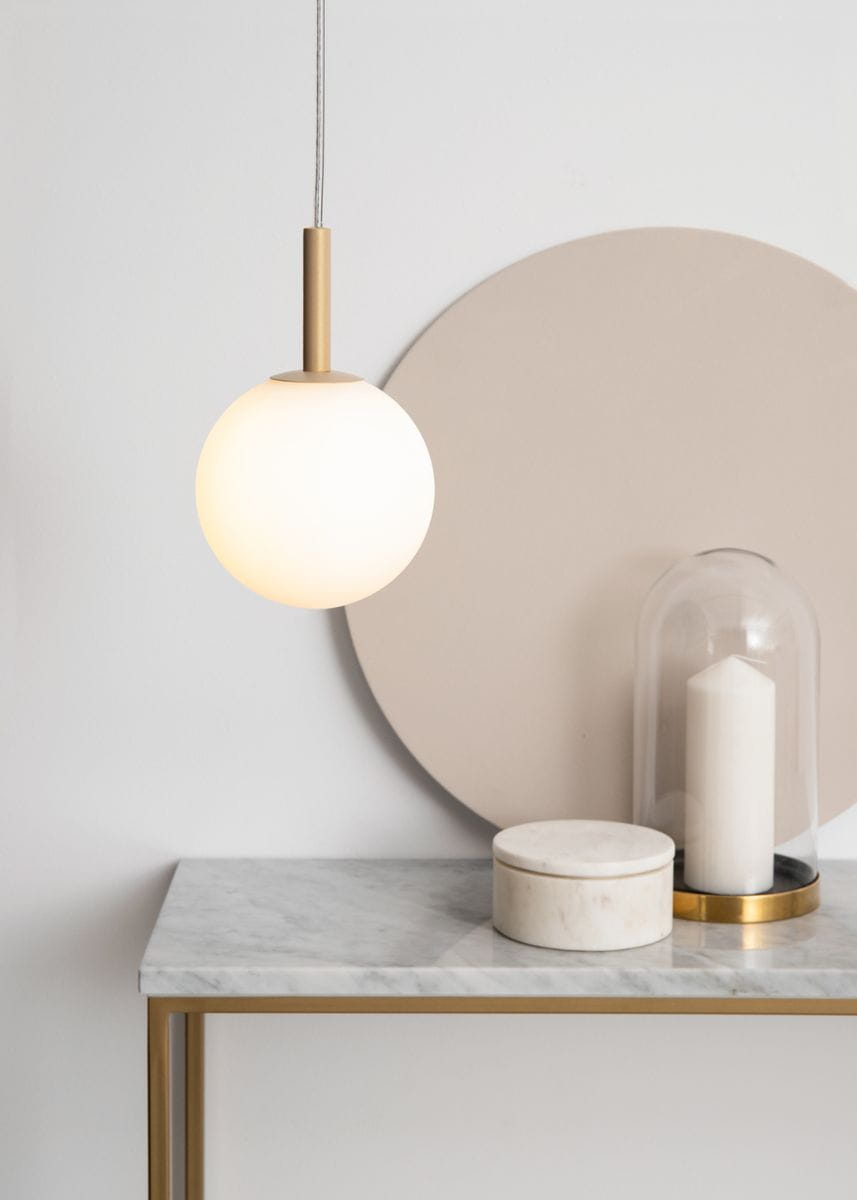 Złota lampa wisząca Gama z kulistym kloszem w kolorze białym, zawieszona na cienkim przewodzie. Ujęcie frontalne, lampa umieszczona nad marmurowym blatem. W tle okrągła dekoracja w odcieniu beżowym.
