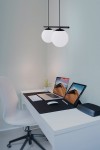 Lampa wisząca Arton 2-punktowa w kolorze czarnym z dwoma okrągłymi kloszami z białego szkła. Zawieszona na cienkich linkach, umieszczona nad biurkiem w jasnym wnętrzu, obok komputerów i rośliny. Proste tło w jasnym kolorze.