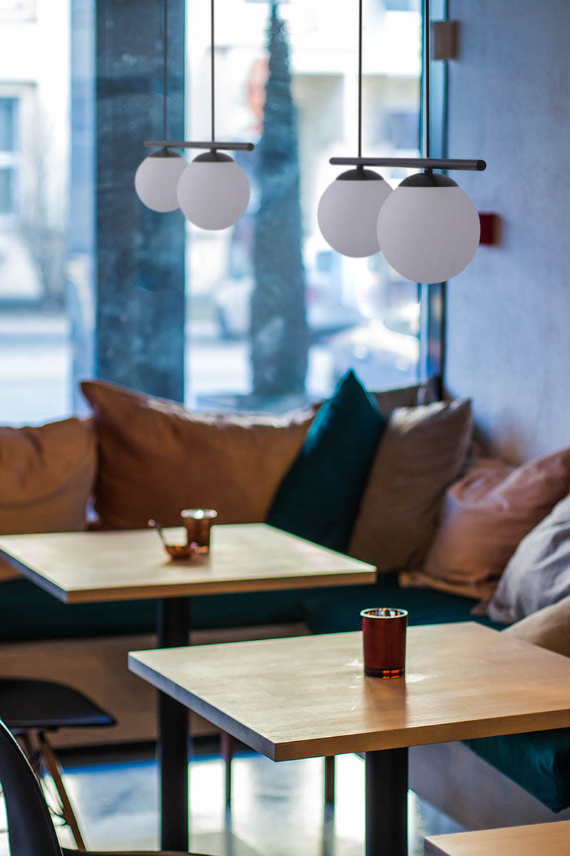 Lampa wisząca Arton 2-punktowa w kolorze czarnym z białymi, okrągłymi kloszami zawieszona w nowoczesnej aranżacji. Widoczna na zdjęciu w kontekście kawiarni z drewnianymi stolikami i miękkimi poduszkami na kanapie.