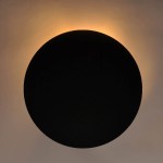 Kinkiet Rega D30 w kolorze czarnym, o okrągłym kształcie, montowany na ścianie. Emituje ciepłe światło wokół krawędzi, tworząc subtelny efekt. Widok z przodu na zamontowany produkt na jasnym tle.