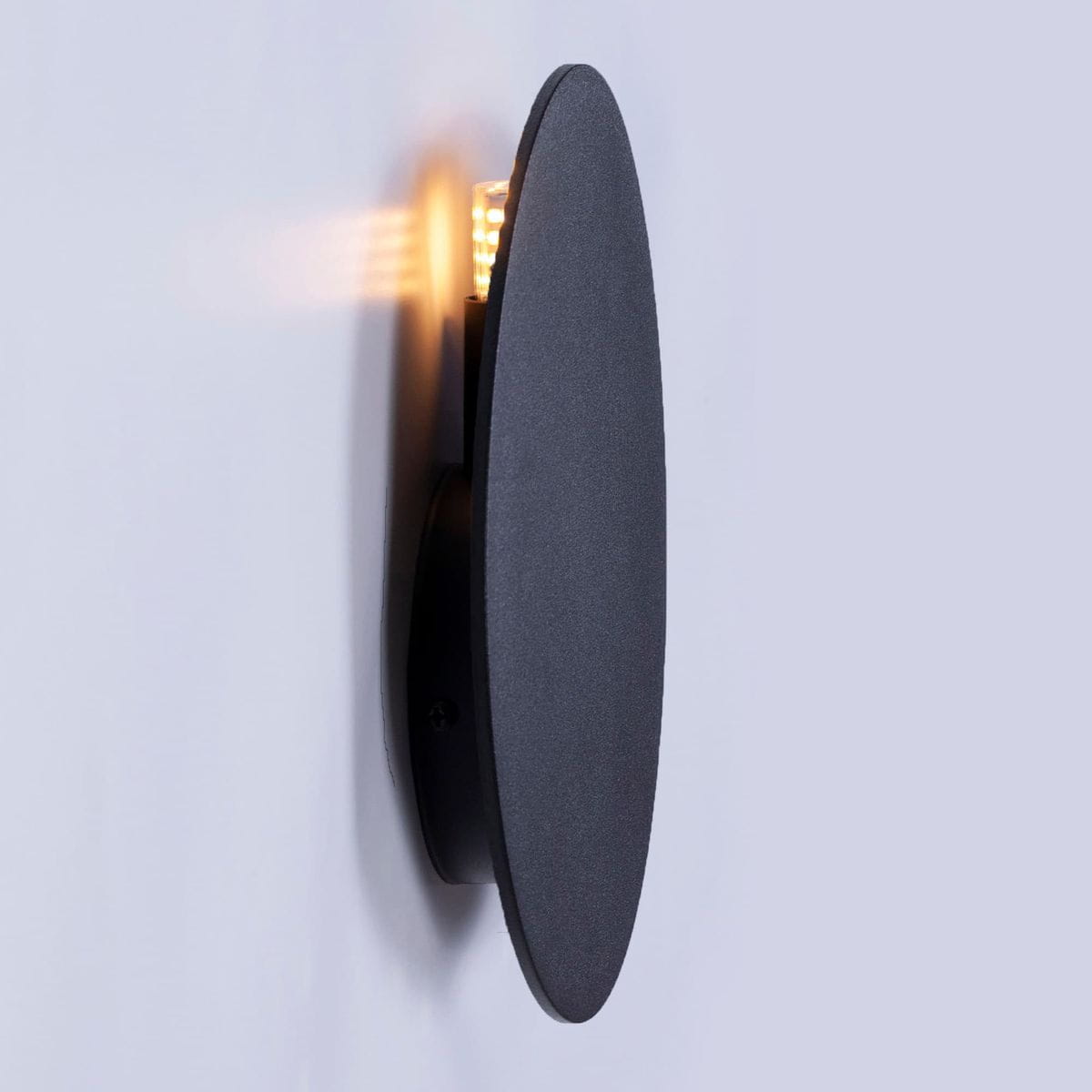 Kinkiet Rega D20 w kolorze czarnym, o owalnym kształcie, zamontowany na ścianie. Z przodu widoczna delikatna iluminacja, która podkreśla jego minimalistyczny design. Produkt leży w poziomie, z matową powierzchnią.