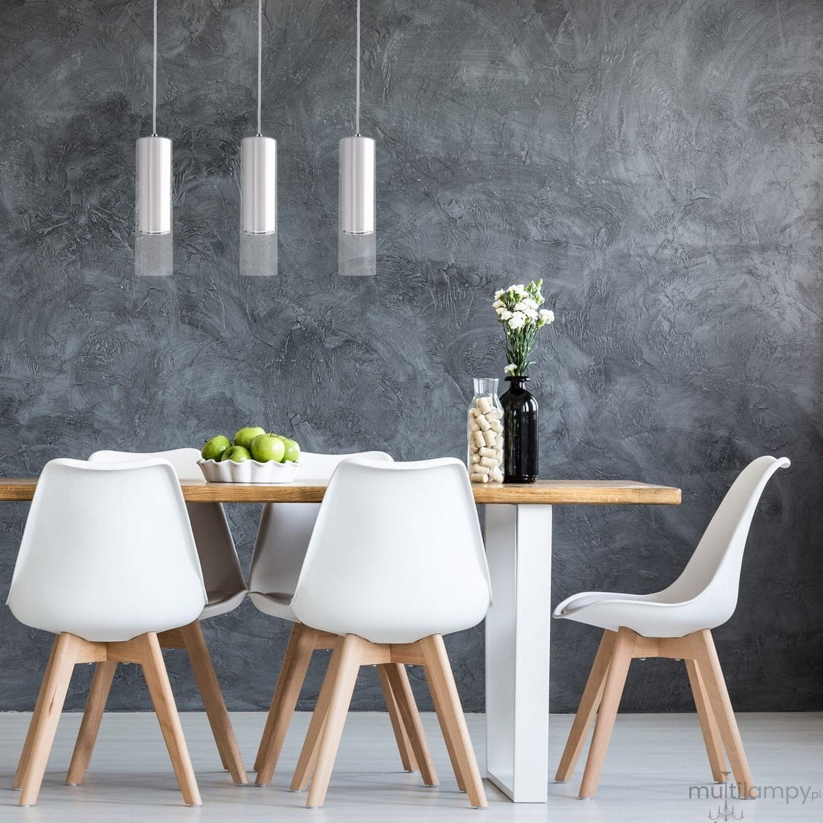 Lampa wisząca Manacor 3-punktowa w białym kolorze, z trzema cylindrycznymi kloszami zawieszonymi na cienkich linkach. Oświetlenie w eleganckim wnętrzu, na tle szarej ściany, nad drewnianym stołem z białymi krzesłami.