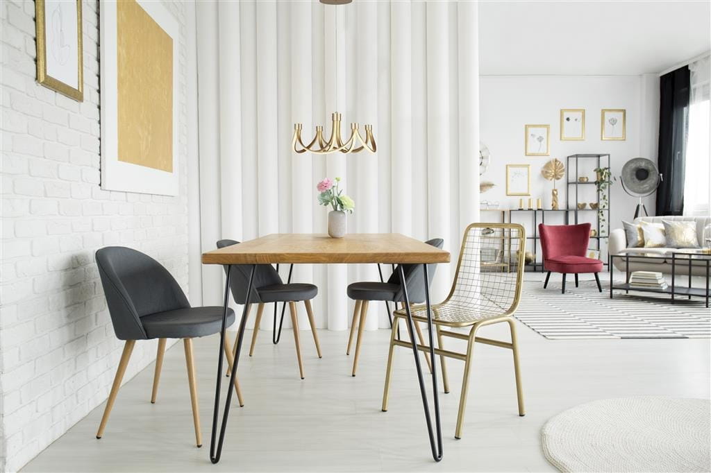 Złota lampa wisząca Signature LED, 8-punktowa, zawieszona nad drewnianym stołem w nowoczesnym wnętrzu. W tle białe ściany, zasłony i różnorodne meble, w tym krzesła i dekoracje. Lampa ma elegancki, minimalistyczny kształt z zaokrąglonymi ramionami.