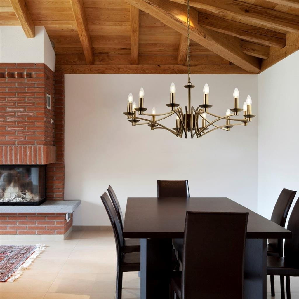 Lampa wisząca Ascot 12-punktowa w kolorze antycznego mosiądzu, z ośmioma ramionami z żarówkami w kształcie świec. Zawieszona nad drewnianym stołem w jadalni, w otoczeniu ciemnych krzeseł i ceglanej kominki.