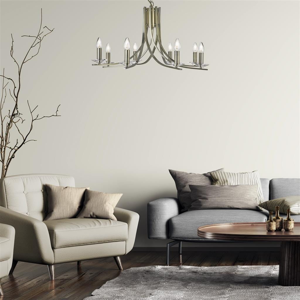Lampa wisząca Ascona 8-punktowa w antycznym mosiądzu, z ośmioma szklanymi kloszami w kształcie świec. Zawieszona w nowoczesnym wnętrzu, na tle jasnej ściany, nad sofą i stolikiem kawowym. Stylowy element oświetleniowy, dodający elegancji pomieszczeniu.