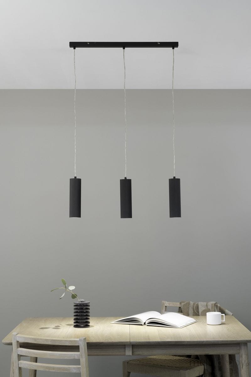 Lampa wisząca Costilla 3-punktowa w kolorze grafitowym. Składa się z trzech cylindrycznych kloszy z rowkowaną fakturą, zawieszonych na cienkich linkach. Montaż sufitowy, umiejscowiona nad drewnianym stołem. Tło w odcieniu szarym.