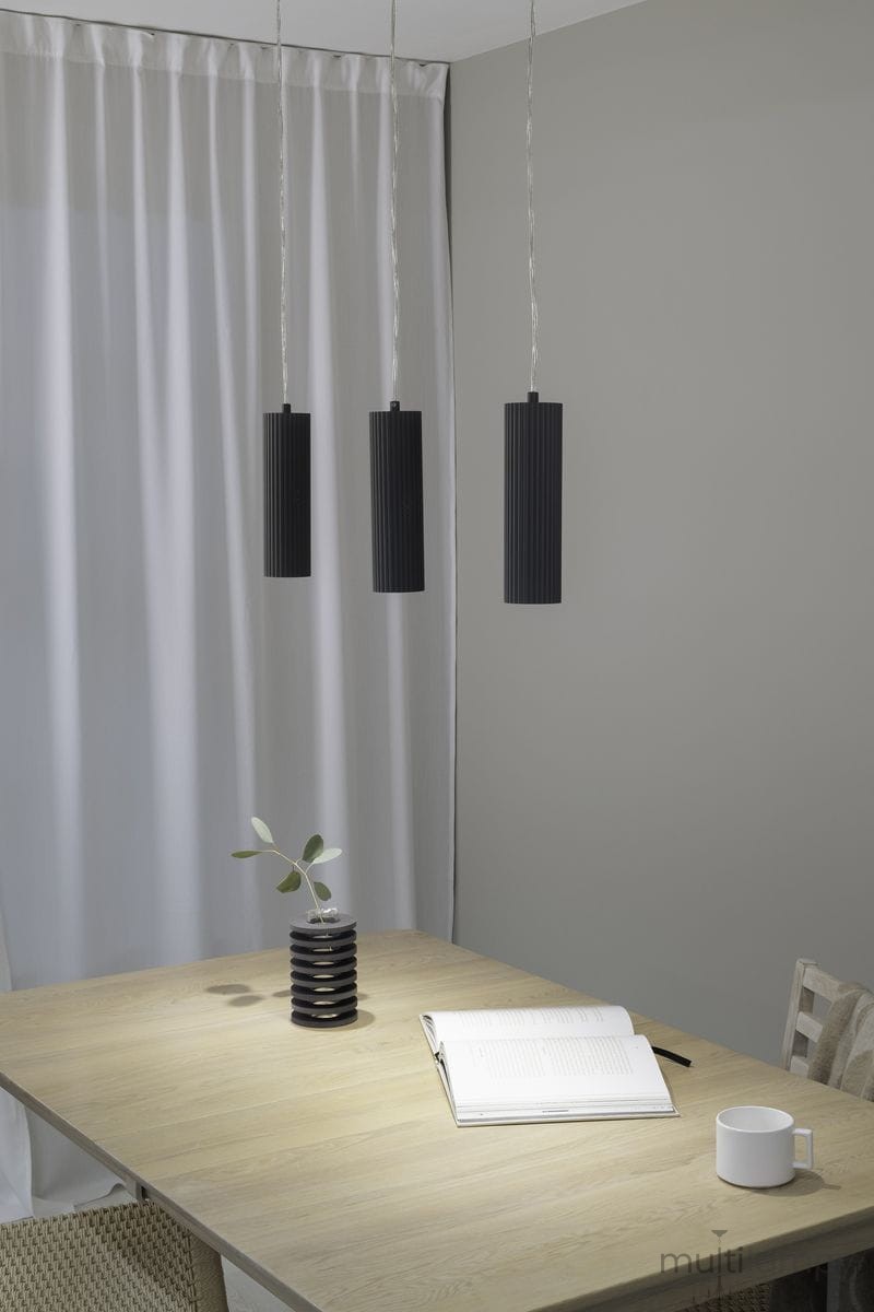 Lampa wisząca Costilla 3-punktowa w kolorze grafitowym z cylindrycznymi kloszami o żebrowanej fakturze, zawieszona na cienkich przewodach. Ujęcie frontalne, w tle stół z drewnianym blatem, notes i filiżanka. Ściana w odcieniu szaro-beżowym.