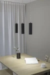 Lampa wisząca Costilla 3-punktowa w kolorze grafitowym z cylindrycznymi kloszami o żebrowanej fakturze, zawieszona na cienkich przewodach. Ujęcie frontalne, w tle stół z drewnianym blatem, notes i filiżanka. Ściana w odcieniu szaro-beżowym.
