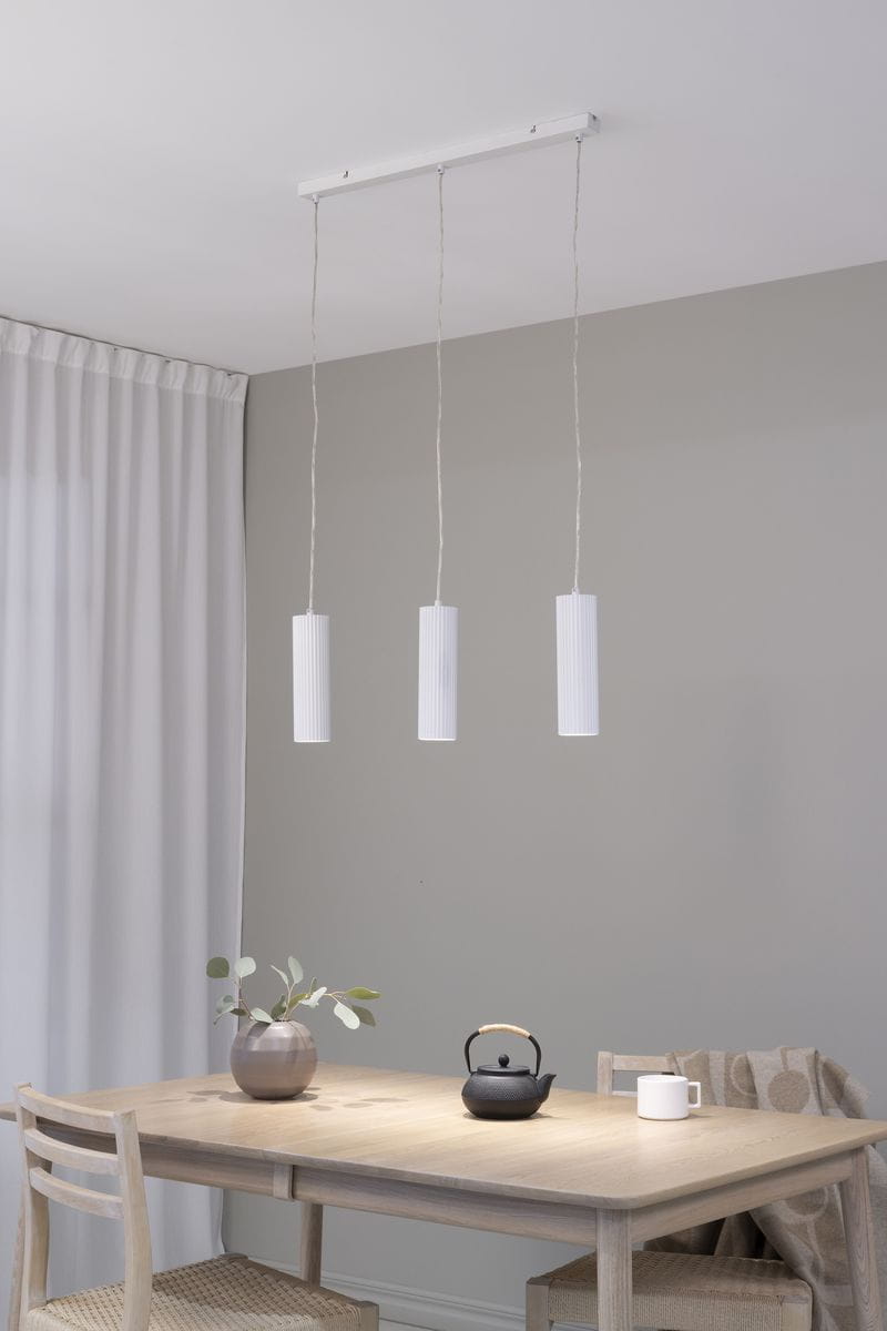 Lampa wisząca Costilla 3-punktowa w kolorze białym, składająca się z trzech cylindrycznych kloszy z teksturą rowków, zawieszona na białym stelażu. Zawieszona nad drewnianym stołem, w tle jasnoszare ściany i zasłony.