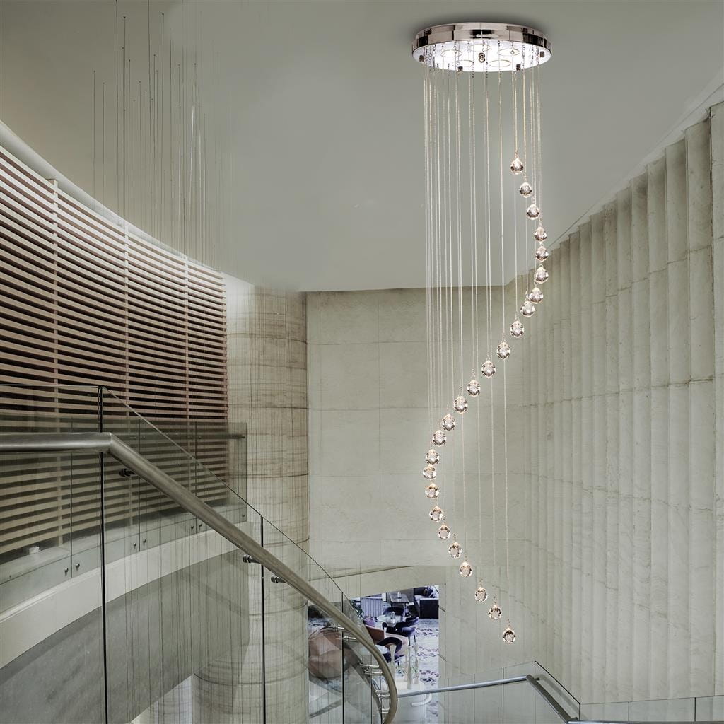 Lampa wisząca 5-punktowa w chromowanym wykończeniu, zawieszona na cienkich linkach. Kształt spiralny z kryształowymi kulkami, oświetlająca przestrzeń nad schodami w nowoczesnym wnętrzu. Surowe, jasne tło podkreśla elegancję lampy.