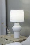 Biała lampa stołowa z abażurem, o okrągłej, żołnierzowatej podstawie i prostym kształcie. Ujęcie z boku, lampa stoi na drewnianym stoliku obok szklanej wazonu z suchymi roślinami. Tło w odcieniach szarości.