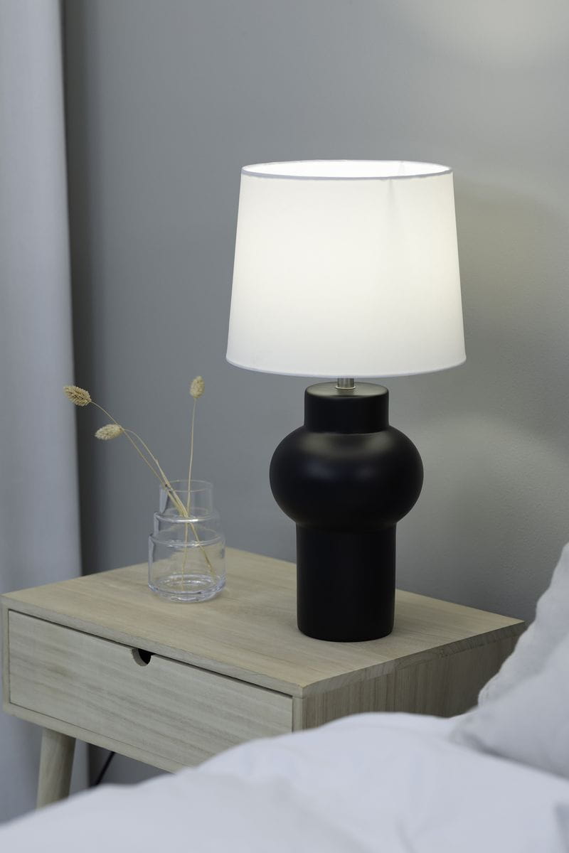 Lampa stołowa Shape w kolorze czarnym z białym abażurem, o okrągłej podstawie i cylindrycznym kształcie, stojąca na drewnianym stoliku nocnym obok przezroczystego wazonu z suszonymi roślinami. Tło w stonowanej kolorystyce.