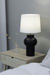 Lampa stołowa Shape w kolorze czarnym z białym abażurem, o okrągłej podstawie i cylindrycznym kształcie, stojąca na drewnianym stoliku nocnym obok przezroczystego wazonu z suszonymi roślinami. Tło w stonowanej kolorystyce.