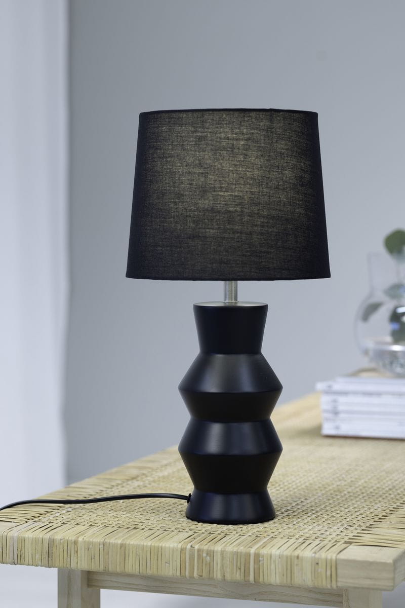 Czarna lampa stołowa Sienna o 1 punkcie świetlnym, stojąca na drewnianym stole. Posiada cylindryczną podstawę z wyraźnymi, falistymi kształtami oraz tekstylny klosz w kolorze czarnym. Widoczny przewód zasilający. Tło jasne, minimalistyczne.
