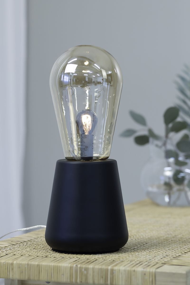 Lampka stołowa Brooke 1-punktowa w czarnej podstawie z matowego materiału, z dużą, przezroczystą żarówką w stylu retro. Ujęcie z boku, na drewnianym stoliku, w tle delikatne rośliny.