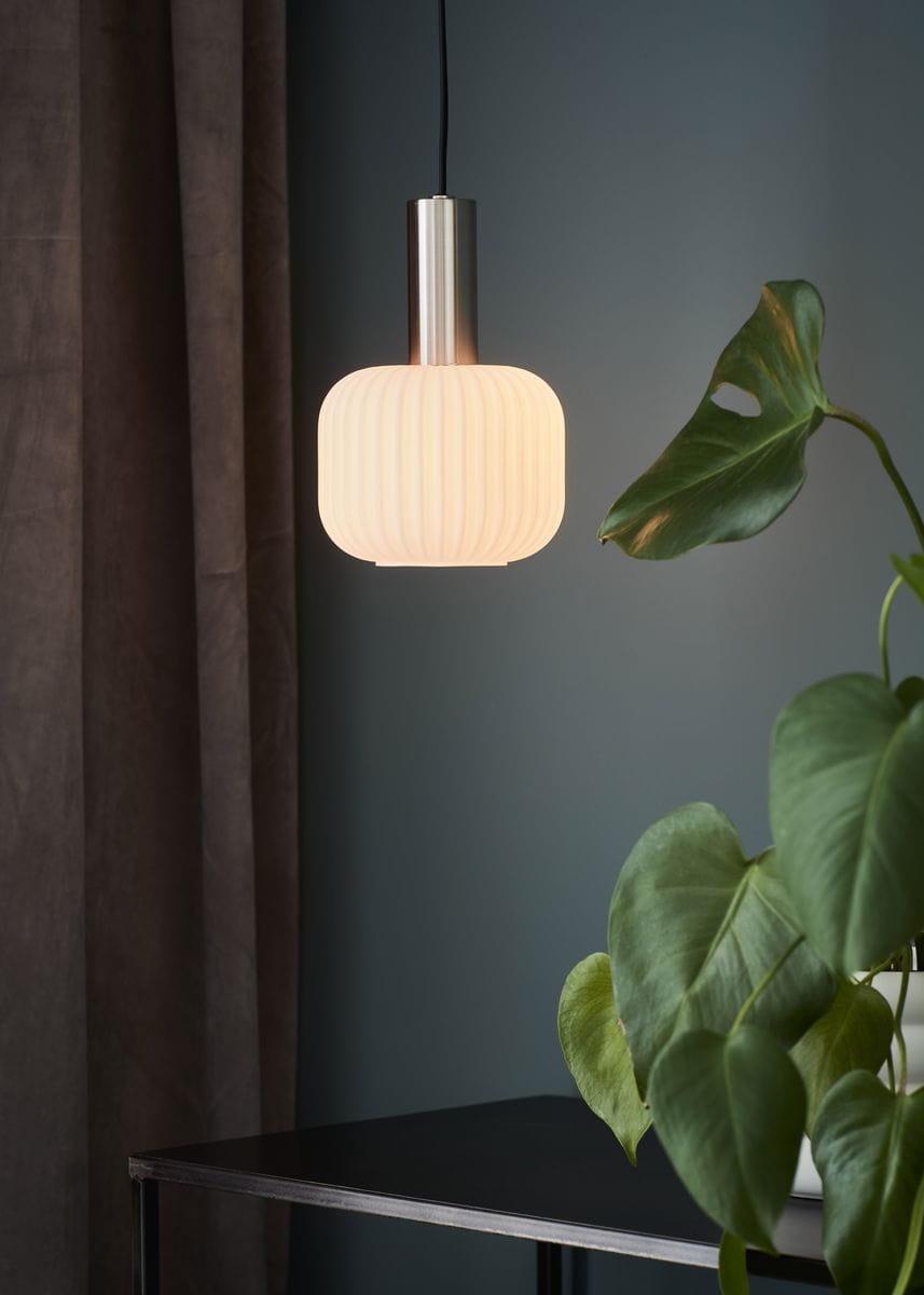 Lampa wisząca Sober D15 w kolorze stalowym i białym, z okrągłym, teksturowanym kloszem. Zawieszona na czarnym przewodzie, w tle zielona roślina i ciemne zasłony. Klosz emituje miękkie światło, nadając przytulny klimat.