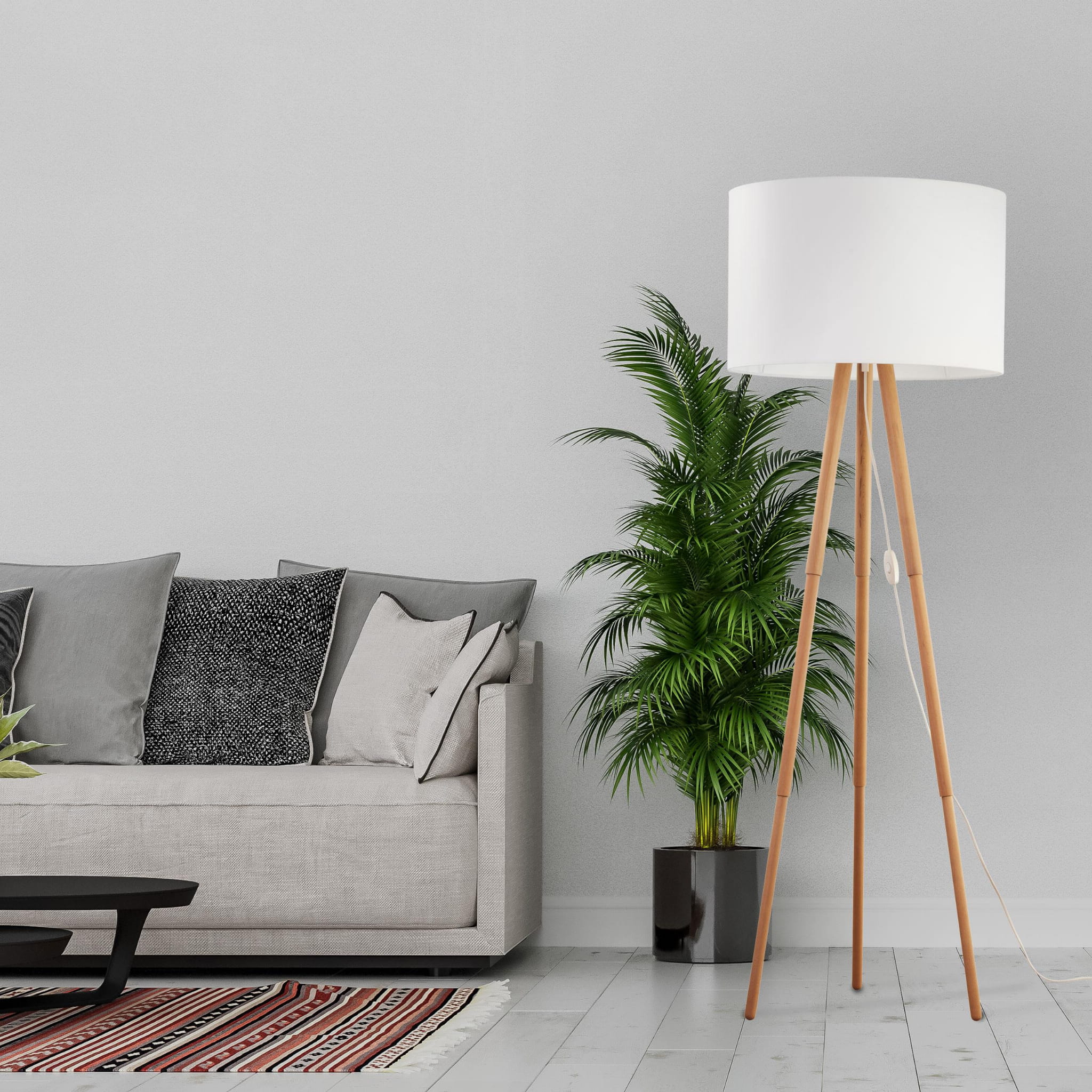Lampa podłogowa Tokyo z białym kloszem na drewnianych nogach z sosny, stojąca w jasnym wnętrzu obok szarej kanapy i rośliny w doniczce. Klosz w kształcie walca, z widocznym włącznikiem na przewodzie.