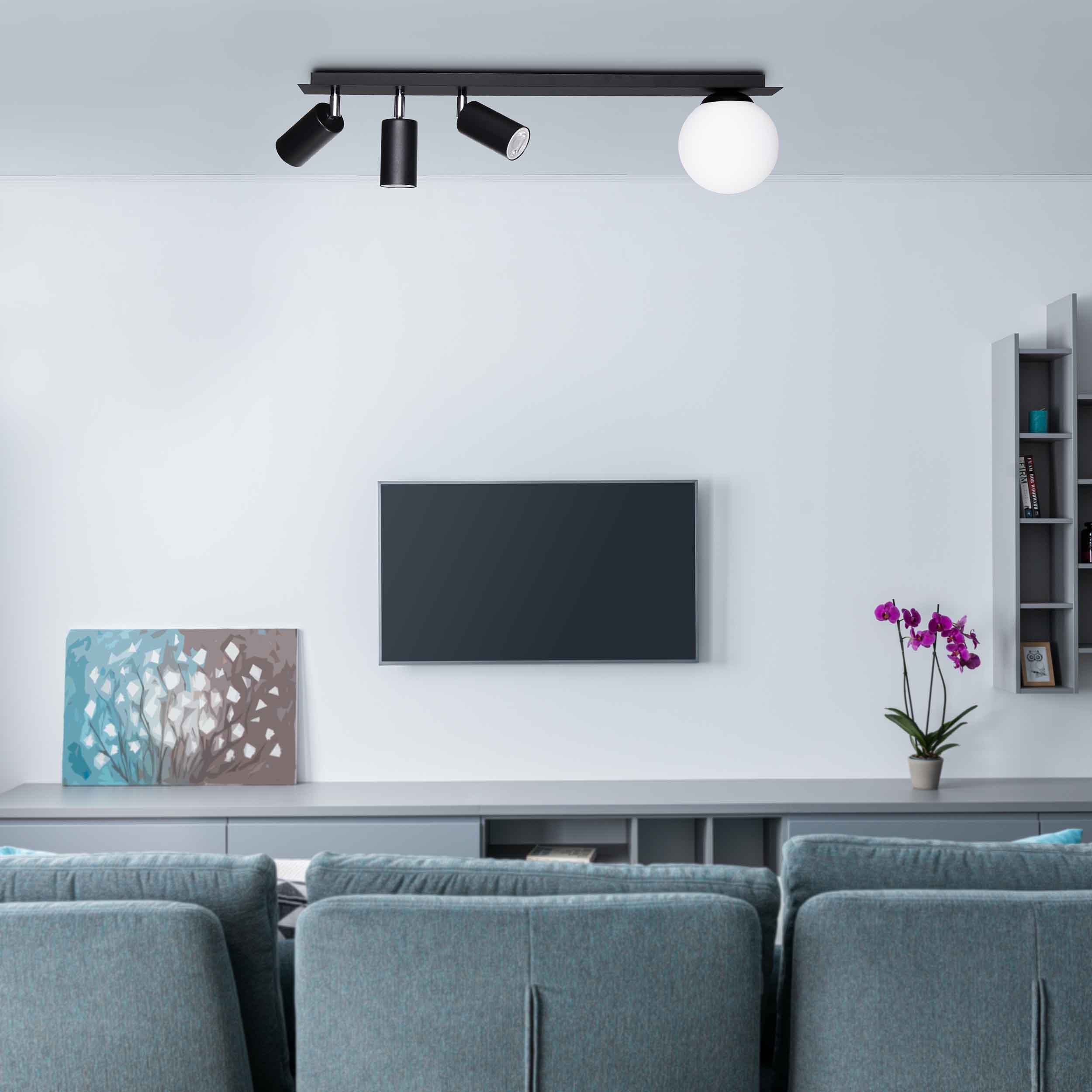 Czarna lampa sufitowa Albio K-4136 z 4 punktami świetlnymi. Dwa reflektory w kształcie cylindrów i jedna okrągła kula. Zawieszona na prostokątnej listwie, w nowoczesnym wnętrzu z szarym telewizorem i kwiatem w doniczce.