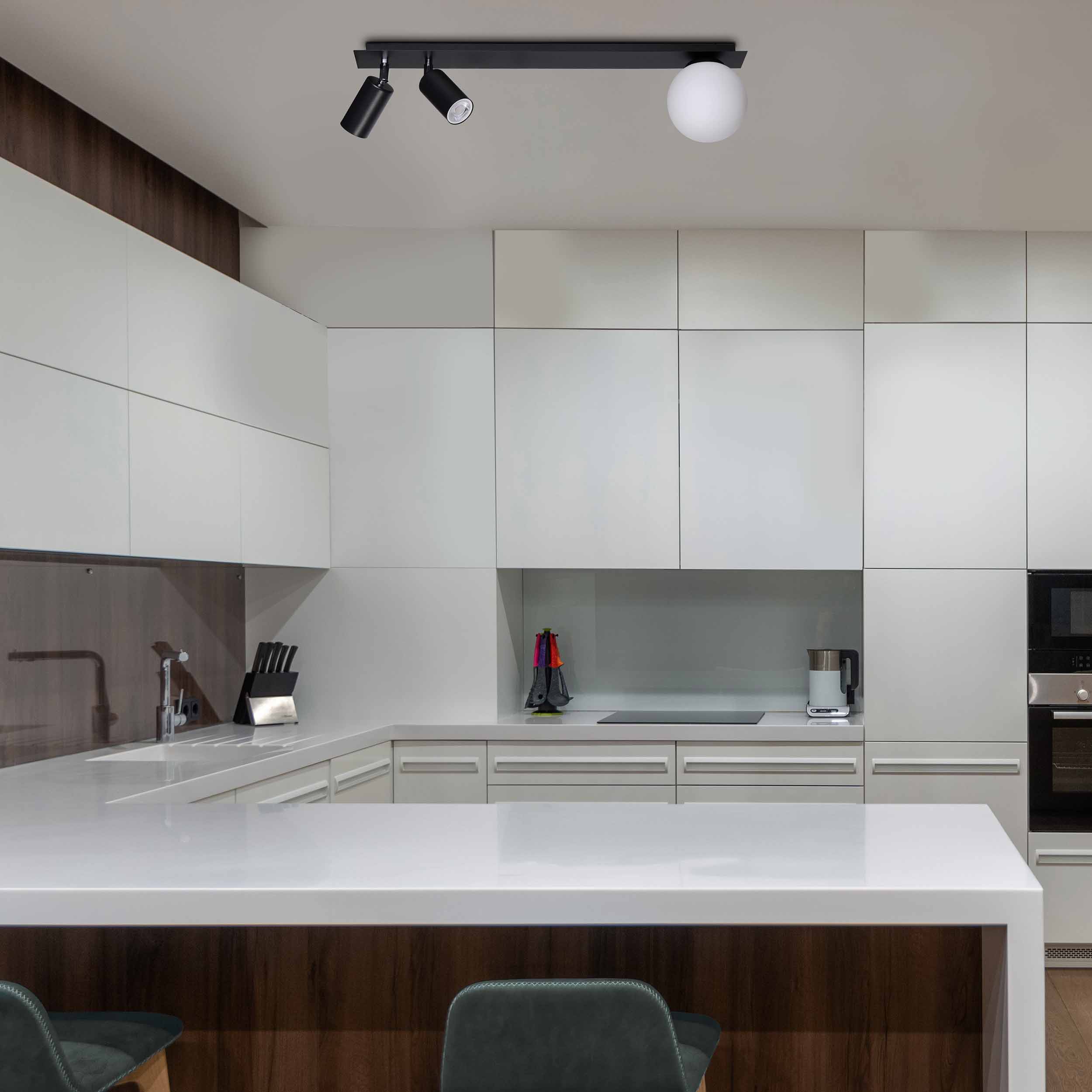 Albio lampa sufitowa 3-punktowa w kolorze czarnym, zamocowana na prostokątnej listwie. Dwa ruchome reflektory oraz jedna okrągła, biała oprawa. Lampa zawieszona w nowoczesnej kuchni z jasnymi szafkami i białym blatem.