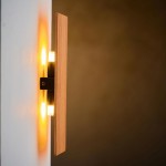Kinkiet Tavola 2-punktowy 3272 w kolorze drewna, z czarnym mocowaniem. Zainstalowany na ścianie, emitujący ciepłe światło. Prosty, minimalistyczny design z jedną drewnianą płytą i dwoma źródłami światła. Tło w odcieniach brązu.