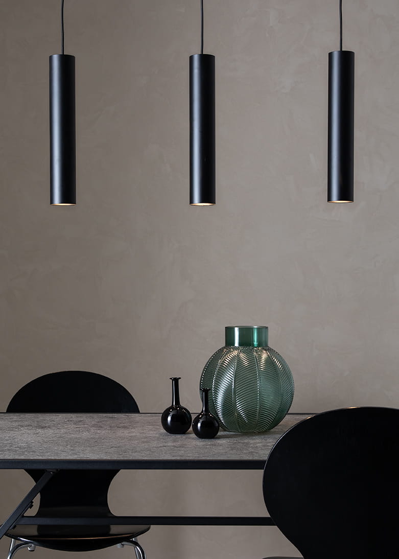 Lampa wisząca 3-punktowa w kolorze czarnym, składająca się z trzech cylindrycznych opraw w równych odstępach, zawieszonych na cienkich linkach. W tle widoczny szklany wazon w odcieniu zieleni oraz czarne dekoracje na stole.