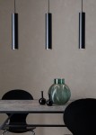 Lampa wisząca 3-punktowa w kolorze czarnym, składająca się z trzech cylindrycznych opraw w równych odstępach, zawieszonych na cienkich linkach. W tle widoczny szklany wazon w odcieniu zieleni oraz czarne dekoracje na stole.