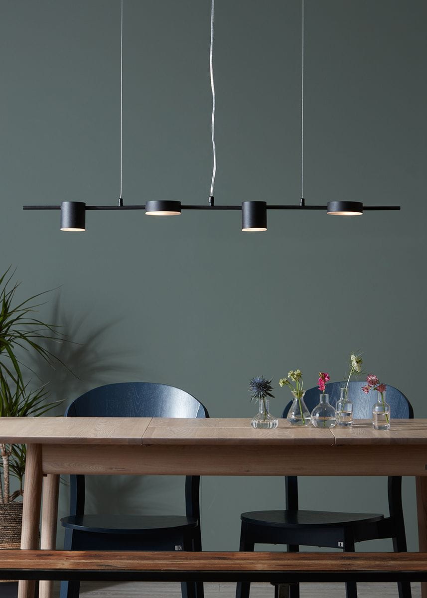 Czarna lampa wisząca 4-punktowa Row, z czterema cylindrycznymi kloszami, zawieszona na cienkim, metalowym pręcie. Ujęcie z przodu, w tle stół z kwiatami w wazonach i krzesła w ciemnym kolorze, na jednolitym, zielonym tle.