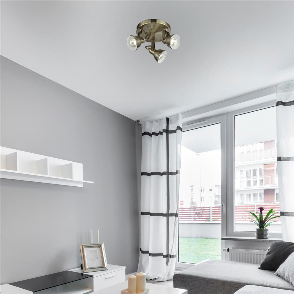 Lampa sufitowa (spot) Focus, 3-punktowa, w kolorze antycznego mosiądzu. Ułożona w sufit, z trzema reflektorami skierowanymi w różnych kierunkach, w nowoczesnym wnętrzu z szarymi ścianami i dużymi oknami.