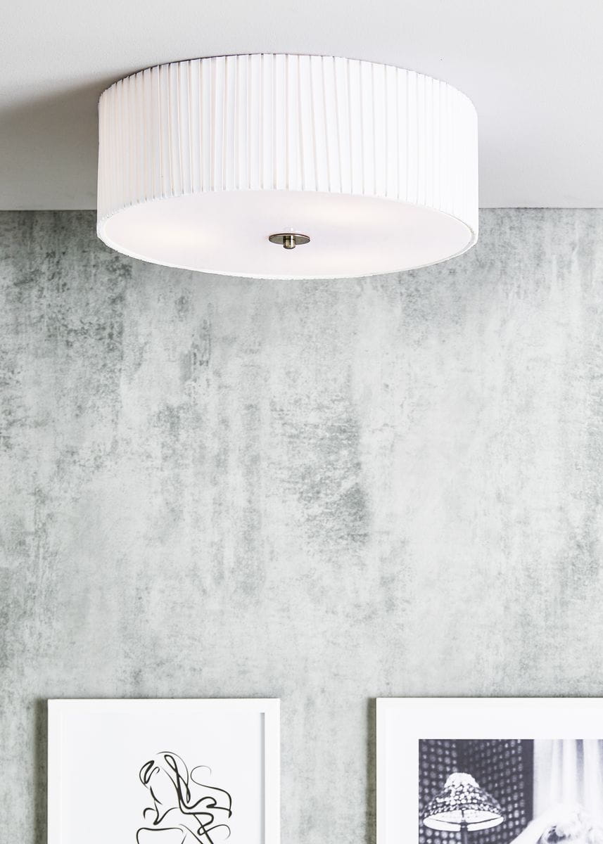 Lampa sufitowa Jackie D45 w kolorze białym, okrągła, z plisowanym kloszem, zamontowana na suficie. Widoczne detale to metalowy element mocujący. Tło w odcieniach szarości z obrazami na ścianie.