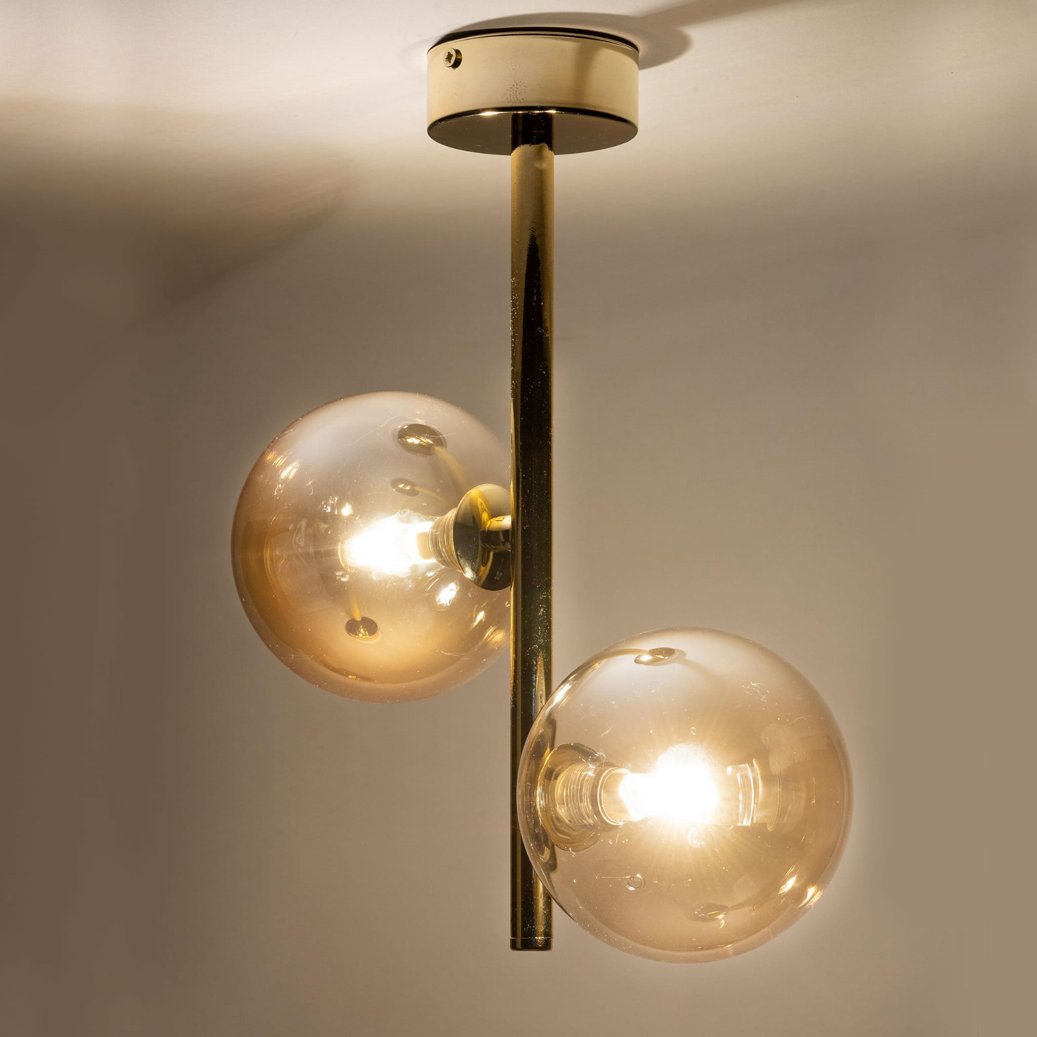 Złota lampa sufitowa Estera z dwoma punktami świetlnymi. Szklane klosze w kształcie kul, umieszczone na pionowym metalowym słupku. Lampa montowana do sufitu, emitująca ciepłe światło. Tło jasne, co podkreśla elegancki design.
