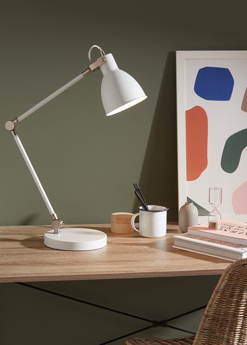 Lampa biurkowa 1-punktowa w kolorze białym i stalowym, z oświetleniem skierowanym w dół. Ustawiona na drewnianym biurku, obok kubka, pojemnika na długopisy i książek. Głowica lampy jest regulowana, a podstawa okrągła i stabilna. Tło w odcieniu zieleni.