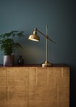 Lampa stołowa Grimstad 1-punktowa w kolorze złotym, z metalowym kloszem i regulowanym ramieniem. Ustawiona na drewnianym blacie z mosiężnymi detalami, obok rośliny w doniczce i dekoracyjnego wazonu. Tło w ciemnym odcieniu.