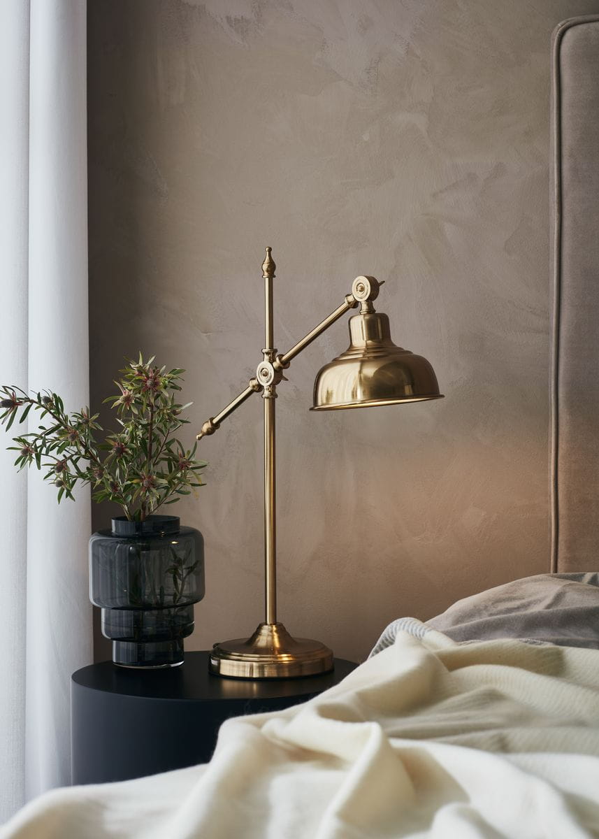 Lampa stołowa Grimstad 1-punktowa w kolorze złotym, z ruchomym ramieniem i kloszem. Ustawiona na czarnym stoliku obok wazonu z rośliną. Tło w odcieniach beżu, z jasno szarym kocem na łóżku.