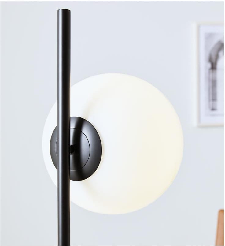 Lampa podłogowa Dione 1-punktowa w kolorze czarnym i białym. Posiada prosty, czarny stelaż oraz okrągły biały klosz. Ujęcie z boku, ukazujące detale mocowania klosza do podstawy. Tło jasne, minimalistyczne.