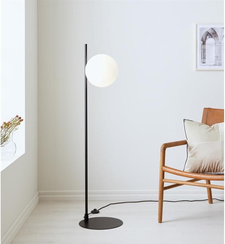 Lampa podłogowa Dione 1-punktowa, czarna z białą kulą, umieszczona w rogu jasnego pomieszczenia. Stojąca na okrągłej podstawie, z cienką, prostą nogą. Obok widoczny drewniany fotel z poduszką. Tło jasne, minimalistyczne.