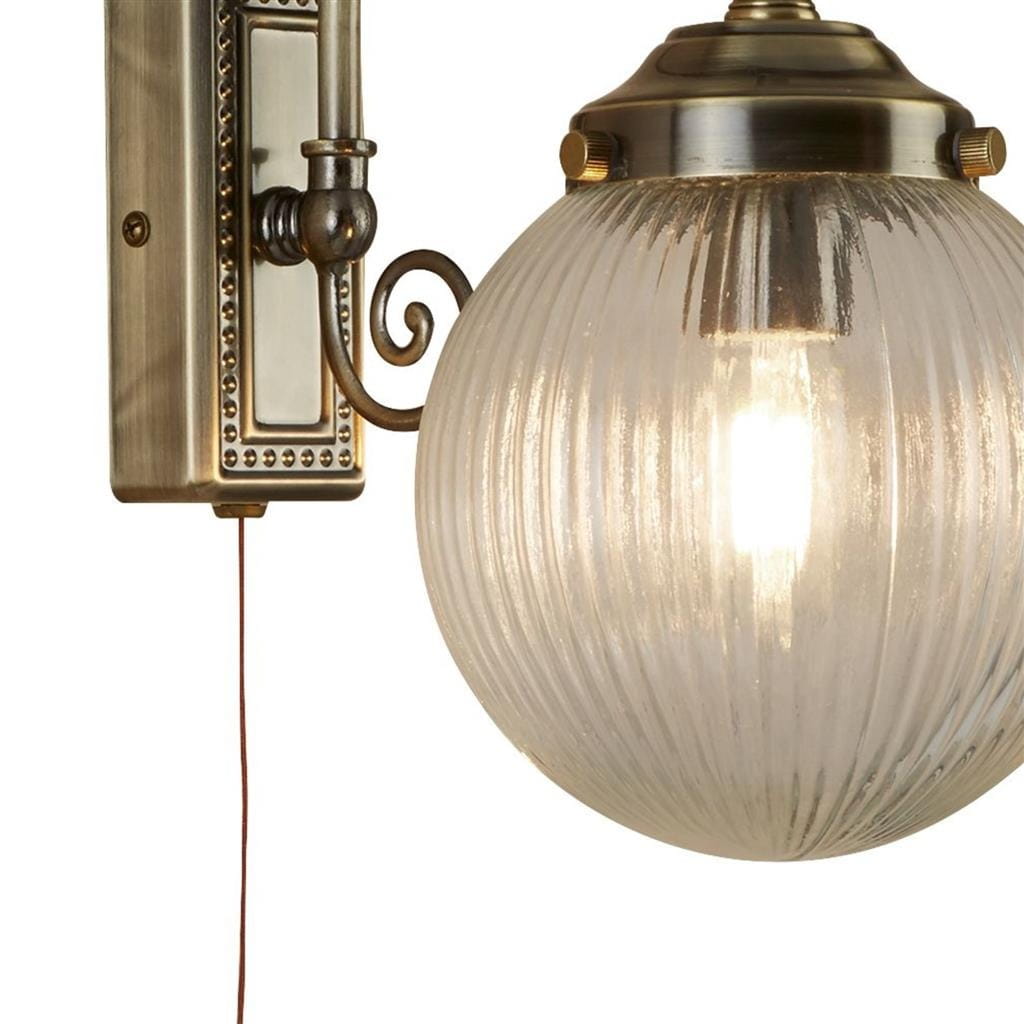 Kinkiet Belvue IP44 w antycznym mosiądzu z przezroczystą, żłobkowaną kulą. Montowany na ścianie, z widocznym przewodem. Okrągła forma lampy, elegancka konstrukcja z dekoracyjnymi detalami.